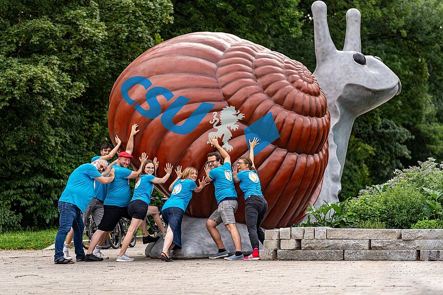 Radentscheid Bayern schiebt CSU-Schnecke an Mehrere Personen des Bündnis Radentscheid Bayern schieben eine große Schnecken-Figur an, auf der "CSU" geschrieben steht.